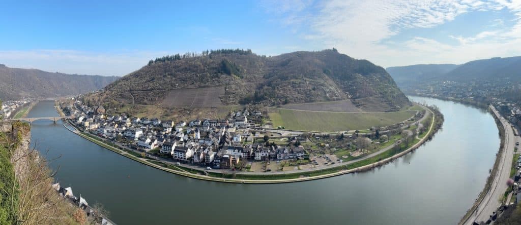 Panorama-Blick von einem Balkon der Reichsburg Cochem auf eine markante Flussschleife der Mosel, die ein kleines Dorf am Fuße bewaldeter Weinberge umschließt.