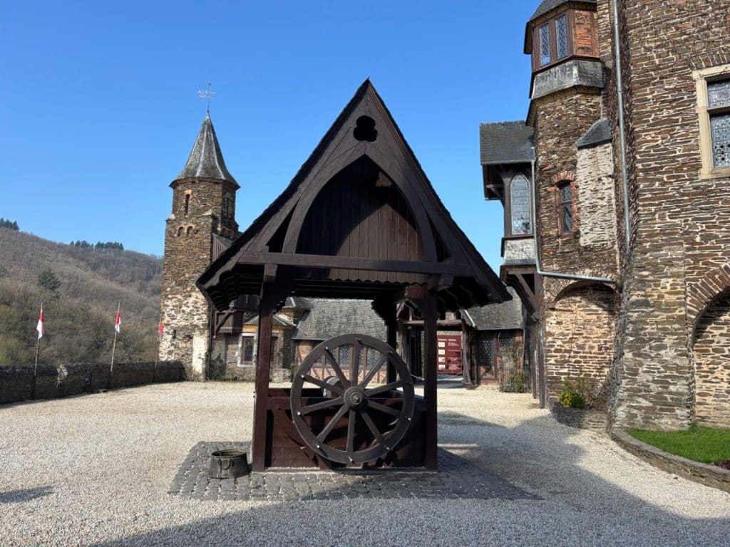 Ein hölzerner Ziehbrunnen mit großem Speichenrad steht im gepflasterten Innenhof der Reichsburg Cochem vor dem Hintergrund historischer Steinmauern und Türme.