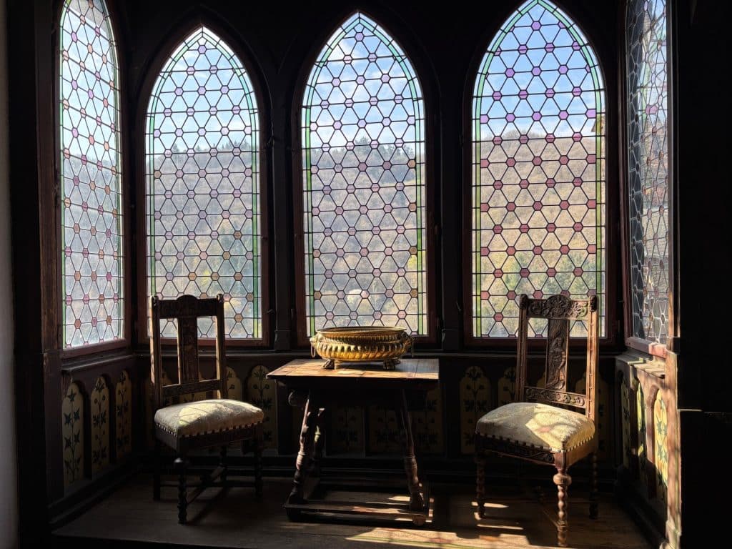 Eine gemütliche Fensternische auf der Reichsburg Cochem mit zwei geschnitzten Holzstühlen, einem kleinen Tisch mit goldener Schale und hohen Spitzbogenfenstern mit bunten Butzenscheiben, die den Blick auf bewaldete Hügel freigeben.