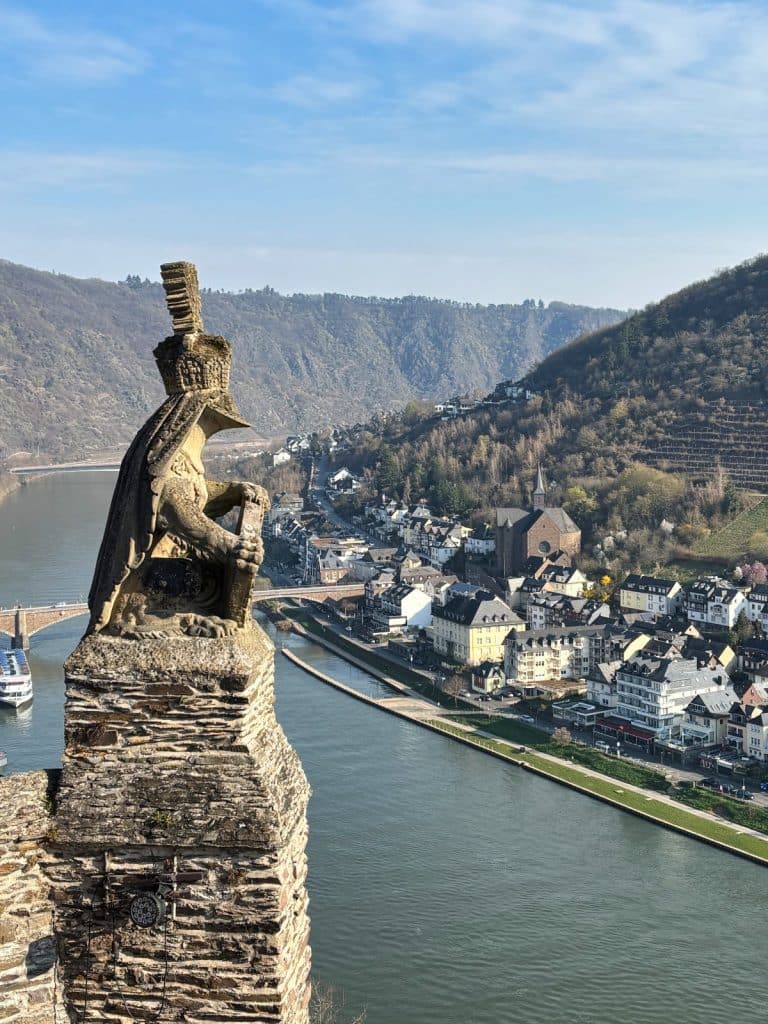 Blick von der Reichsburg Cochem auf eine steinerne Löwenstatue im Vordergrund, die über die Mosel und die historische Altstadt von Cochem wacht.