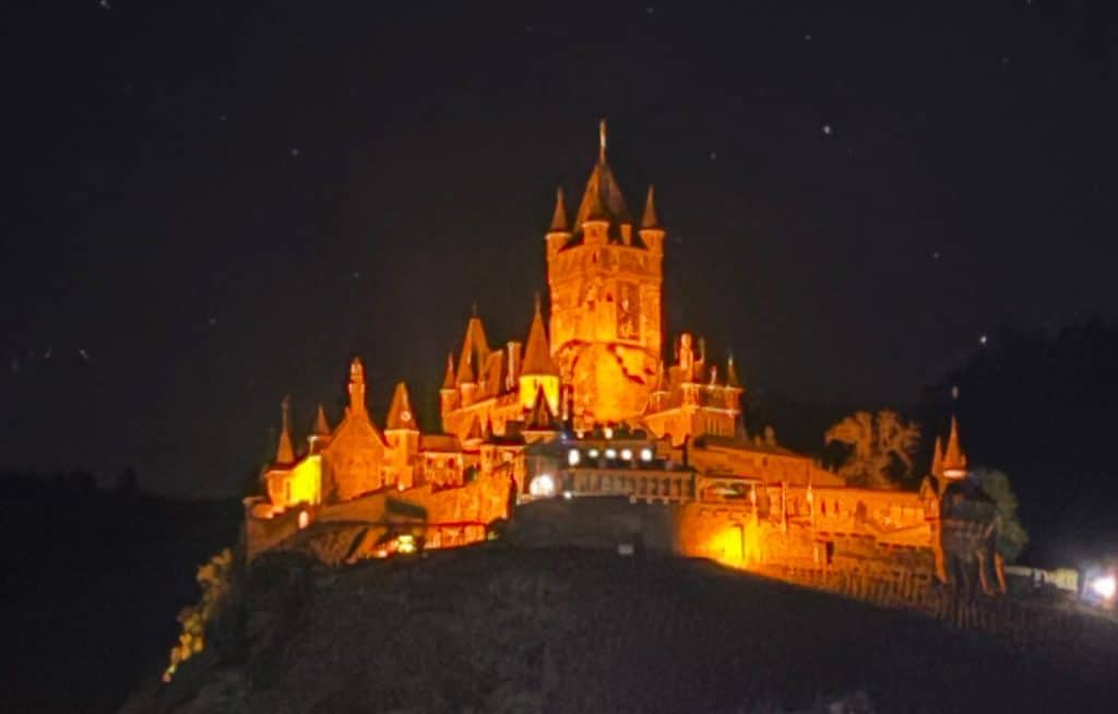 Die Reichsburg Cochem erstrahlt bei Nacht in warmem, orangefarbenem Licht vor einem dunklen Sternenhimmel auf einem Hügel.
