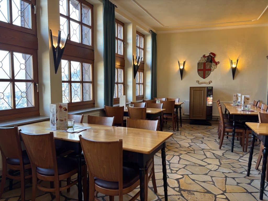 Ein rustikaler Gastraum im Restaurant der Reichsburg Cochem mit Holztischen, Stühlen und großen Sprossenfenstern, die viel Tageslicht hereinlassen. An der hellen Wand hängt ein historisches Wappen zwischen zwei Fackelleuchten über einem unregelmäßigen Natursteinboden.