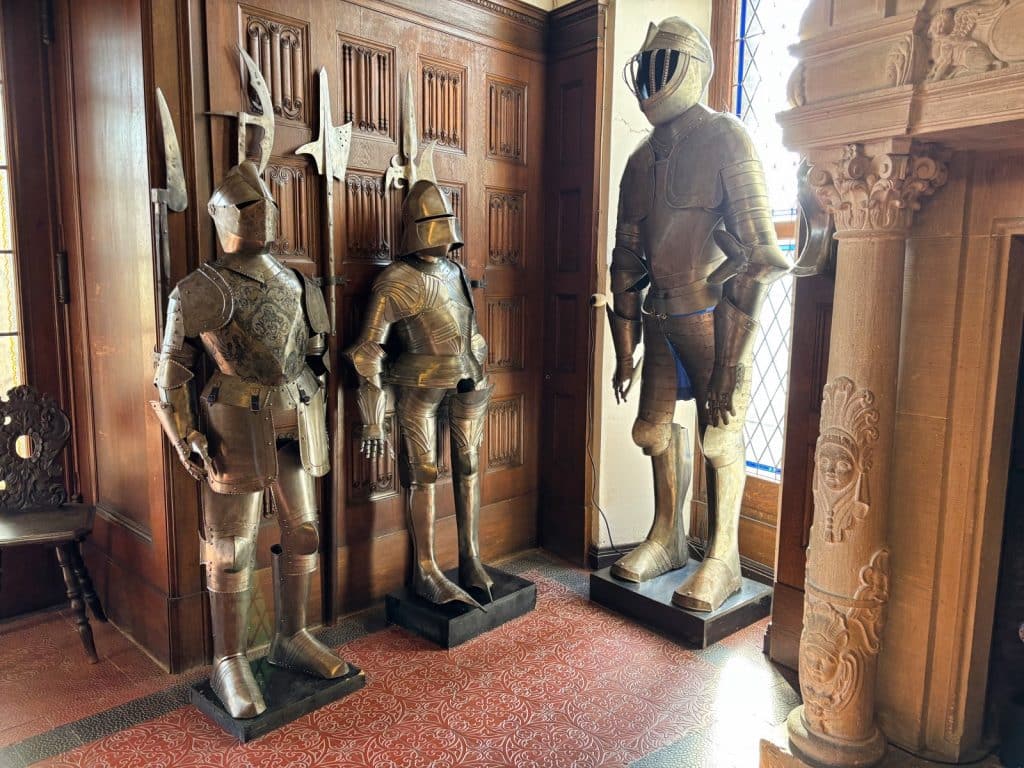 Drei historische Ritterrüstungen stehen auf Podesten vor einer holzgetäfelten Wand in der Reichsburg Cochem, wobei im Hintergrund verschiedene Hellebarden und Waffen zu sehen sind.