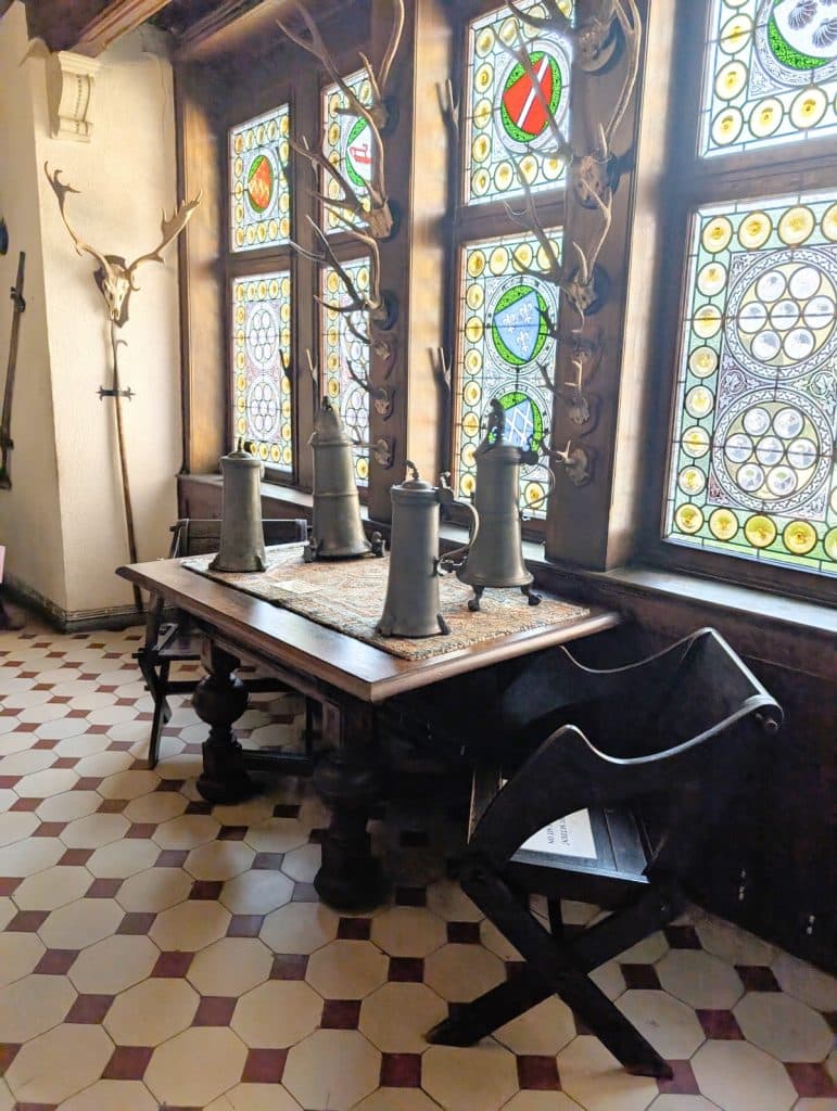 Vier historische Zinnkrüge stehen auf einem rustikalen Holztisch in der Reichsburg Cochem, umgeben von dunklen Holzstühlen, Jagdtrophäen an den Wänden und farbenfrohen Bleiglasfenstern.