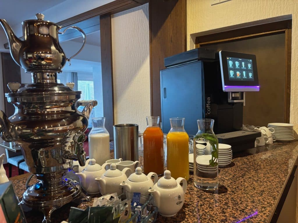 Ein reichhaltiges Getränkebuffet zum Frühstück in einem Hotel in Cochem mit einem glänzenden Samowar, verschiedenen Säften in Glaskaraffen, kleinen weißen Teekannen und einem modernen Kaffeevollautomaten.