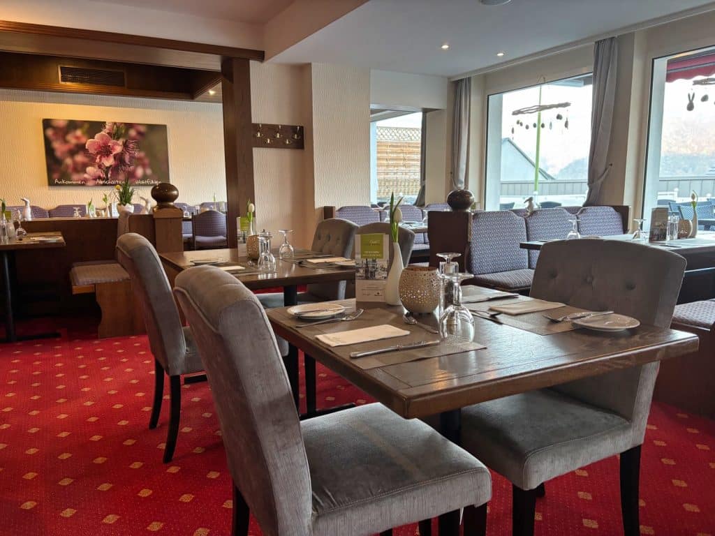 Ein gemütlich eingerichtetes Restaurant im Hotel in Cochem mit eingedeckten Holztischen, grauen Polsterstühlen und einem roten Teppichboden vor großen Fensterfronten.