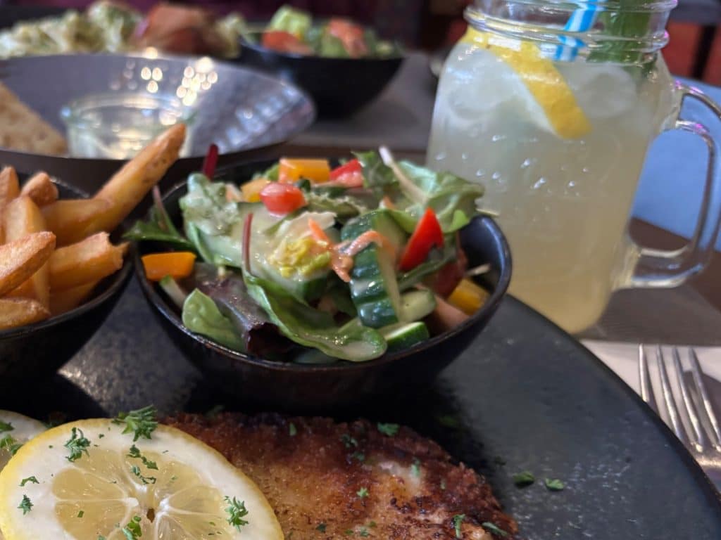 Ein gedeckter Tisch im Restaurant Herbs im Hotel Hegenbarth's mit einem panierten Schnitzel mit Zitronenscheibe, Pommes frites, einem Beilagensalat und einer hausgemachten Limonade im Henkelglas.