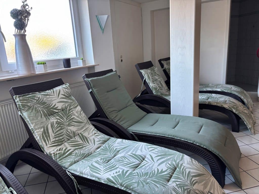 Ein heller Ruheraum im Hotel Hegenbarth's mit mehreren dunklen Rattan-Liegen, die mit grün gemusterten Palmenblatt-Auflagen und einfarbig grünen Polstern ausgestattet sind.
