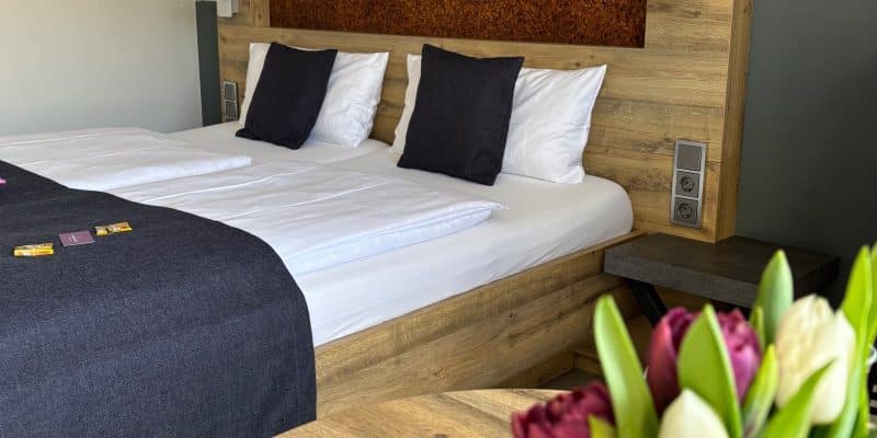 Ein modern eingerichtetes Hotelzimmer in Cochem mit einem großen Doppelbett aus hellem Holz, dunklen Akzentkissen und einem Blumenstrauß aus Tulpen im Vordergrund.