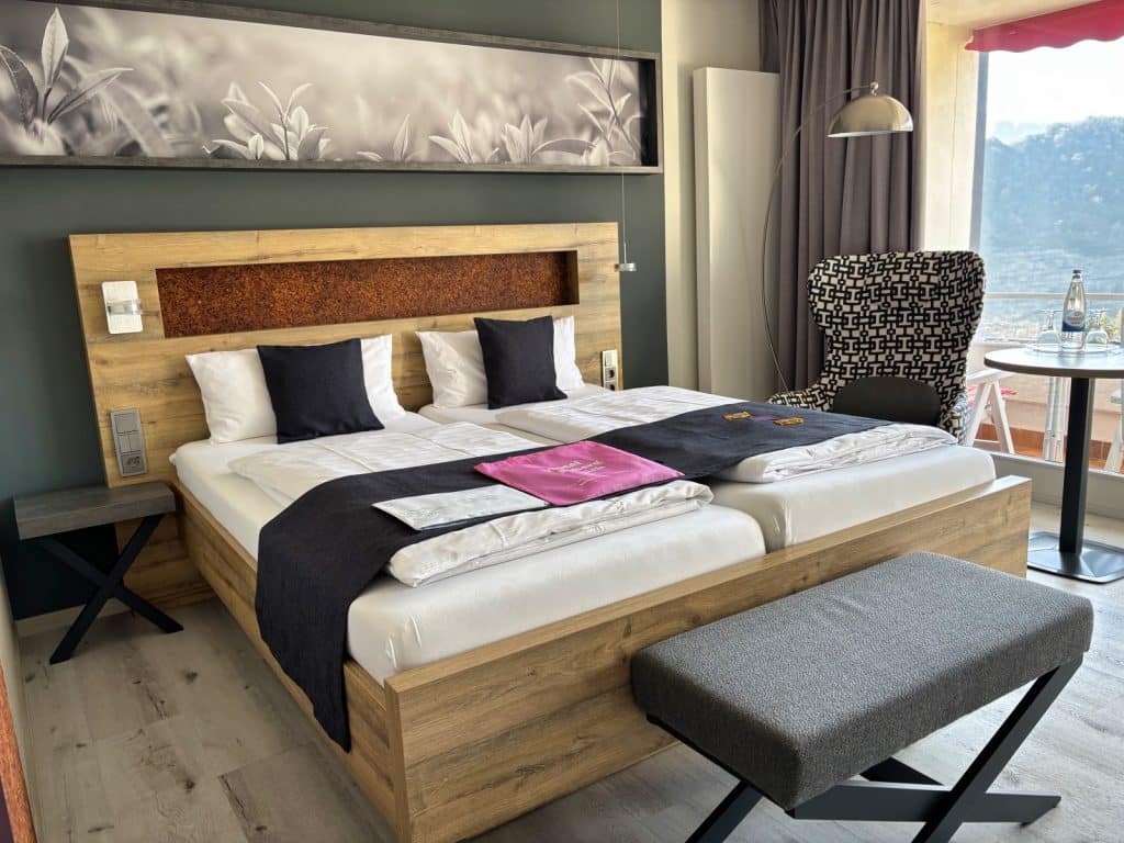 Ein modern eingerichtetes Hotelzimmer in Cochem mit einem großen Doppelbett aus Holz, einer grauen Polsterbank am Fußende und einem Sessel vor einem Fenster mit Ausblick.
