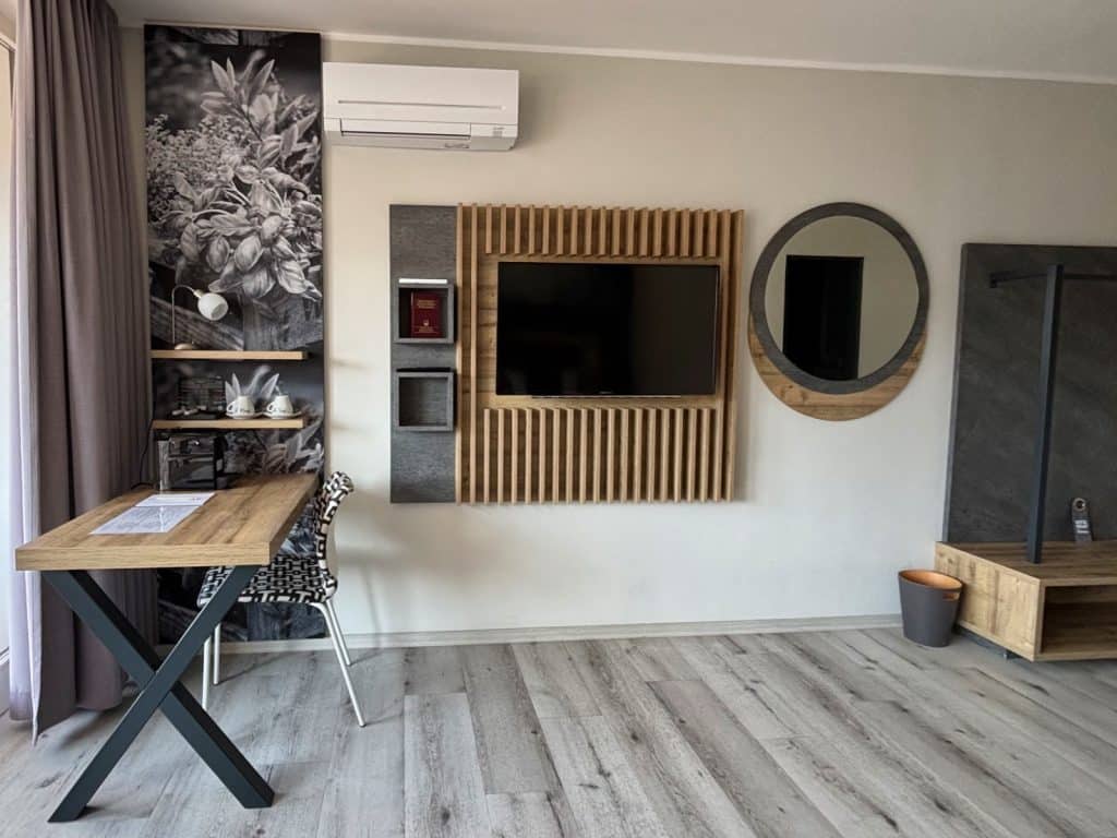 Ein modern eingerichtetes Hotelzimmer in Cochem mit einem hölzernen Schreibtisch, einem Wandfernseher auf einer Lamellenwand und einem runden Spiegel über hellem Laminatboden.