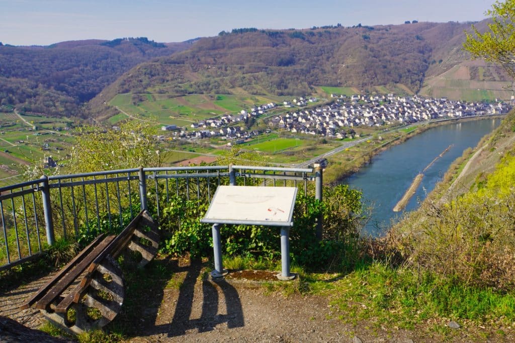 Blick von einem Aussichtspunkt mit Bank und Informationstafel auf das Moselland mit einer Flussschleife der Mosel, einem Dorf und bewaldeten Hängen.