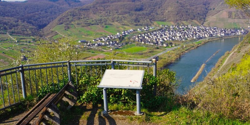 Blick von einem Aussichtspunkt mit Bank und Informationstafel auf das Moselland mit einer Flussschleife der Mosel, einem Dorf und bewaldeten Hängen.