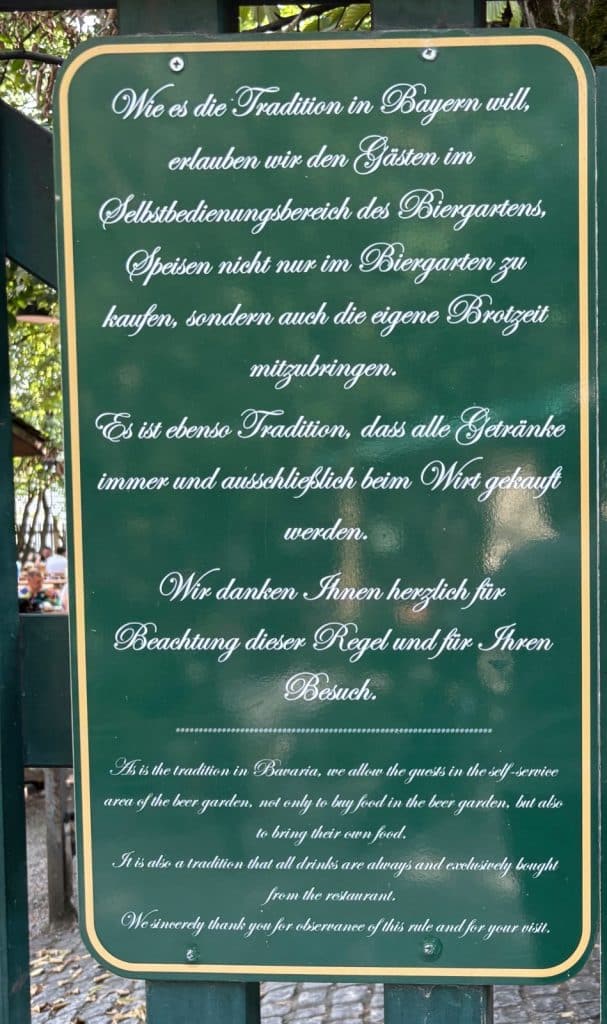 Ein grünes Hinweisschild im Augustiner-Keller in München erklärt in weißer Schreibschrift auf Deutsch und Englisch die bayerische Biergartentradition, nach der eigene Speisen mitgebracht werden dürfen, Getränke jedoch vor Ort gekauft werden müssen.