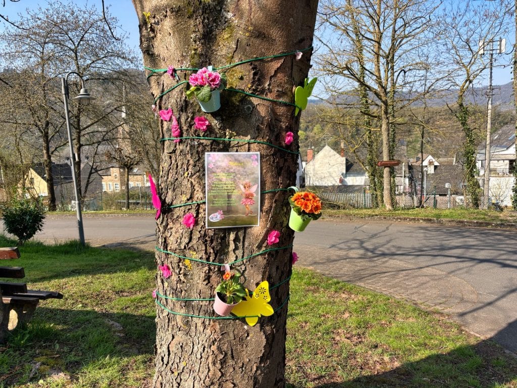 Ein mit bunten Blumenampeln, Schmetterlingen und einem Märchenbild geschmückter Baumstamm am Wegrand der Neef-Wanderung für Kinder.
