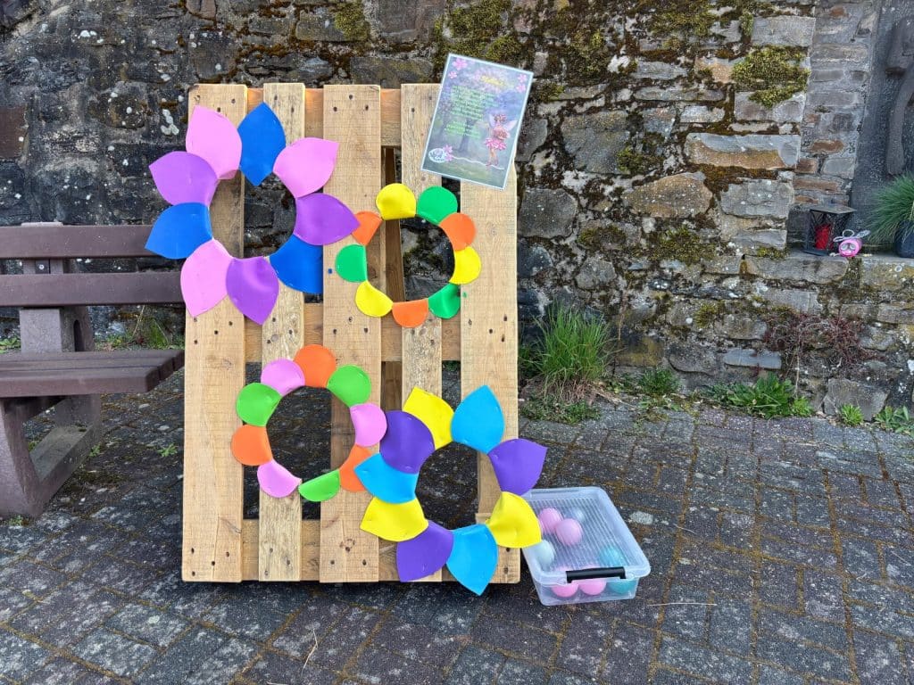 Eine hölzerne Palette dient als Kinderstation auf einer Wanderung bei Neef und zeigt vier bunte, aus Kunststoffblättern geformte Blumen mit Wurflöchern sowie eine Kiste mit kleinen Bällen davor.