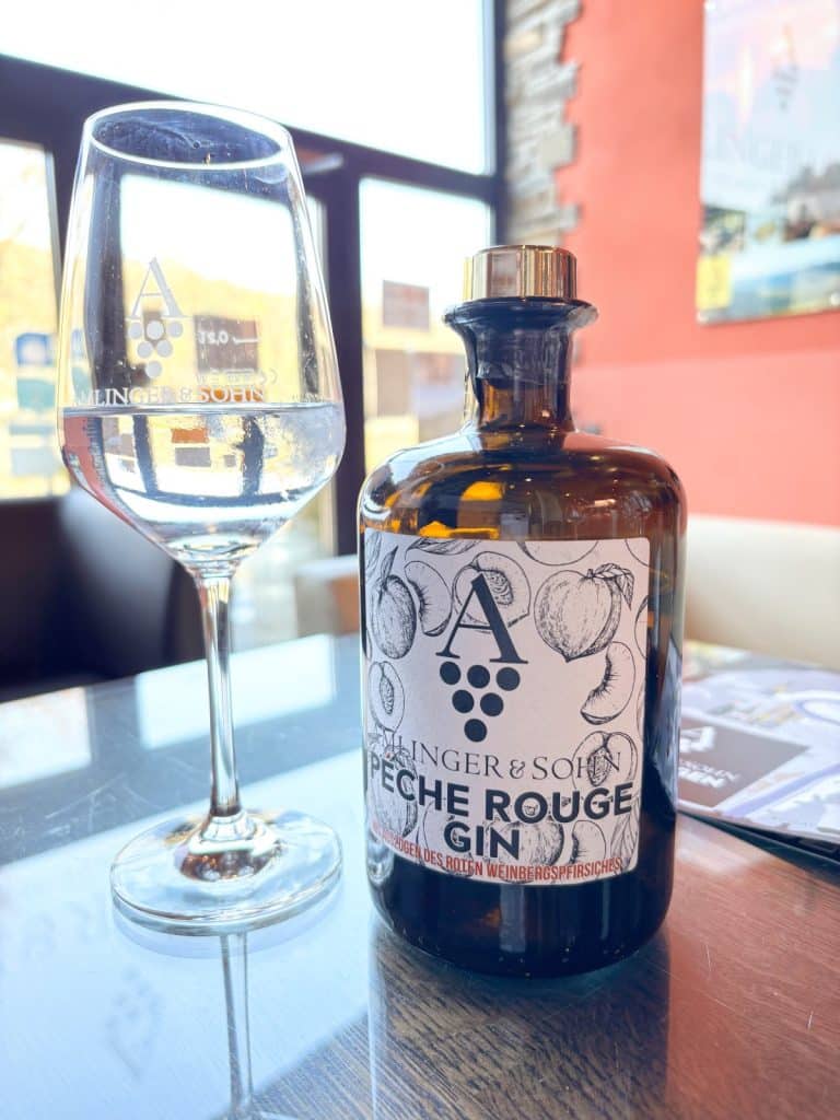 Eine Flasche „Peche Rouge Gin“ des Weinguts Amlinger & Sohn steht neben einem gefüllten Weinglas auf einem glänzenden Tisch im Neef Weingut.