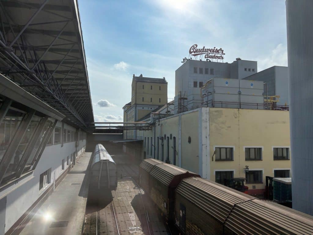 Blick über das Betriebsgelände der Budweiser Budvar Brauerei mit Güterwaggons auf Schienenwegen zwischen Lagergebäuden und dem markanten Budweiser-Schriftzug auf einem Dach.