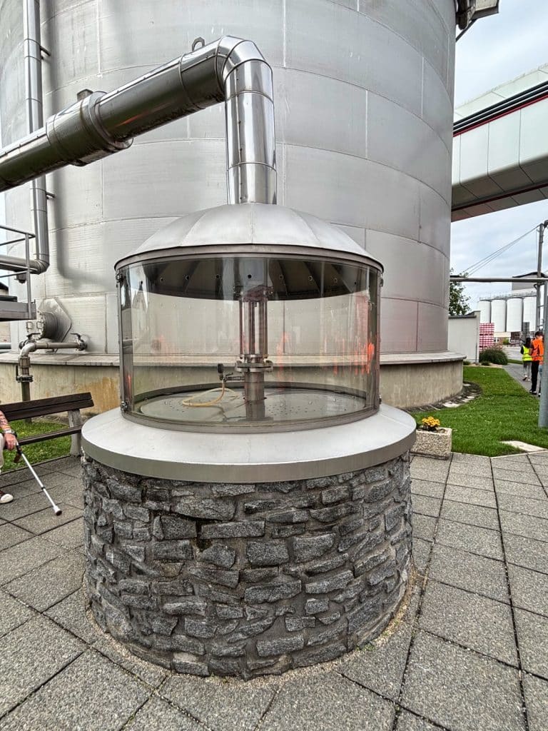 Ein gläserner Brunnen mit einer Steinfassung und einer metallenen Kuppel steht im Außenbereich der Budweiser Brauerei vor einem großen silbernen Silo. Eine dicke Metallleitung führt von oben in den Brunnen, der Teil einer Brauereiführung ist.