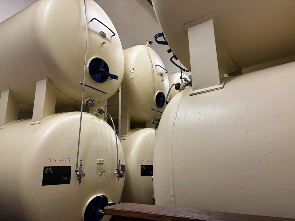 Große, beige Lagertanks in der Budweiser Brauerei sind in einem Kellerraum übereinandergestapelt und mit verschiedenen Ventilen, Leitungen und handschriftlichen Markierungen versehen.