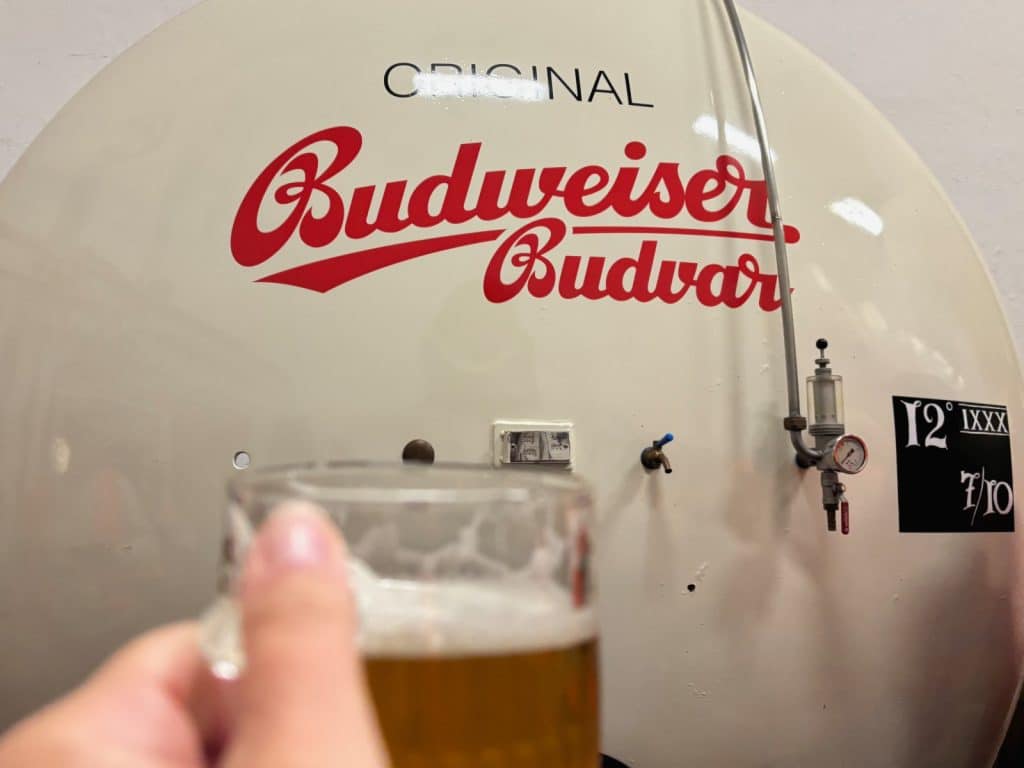 Eine Hand hält bei einer Brauereiführung in Budweis ein frisch gezapftes Glas Bier zum Anstoßen vor einen großen, weißen Lagertank mit dem roten Logo von Budweiser Budvar.