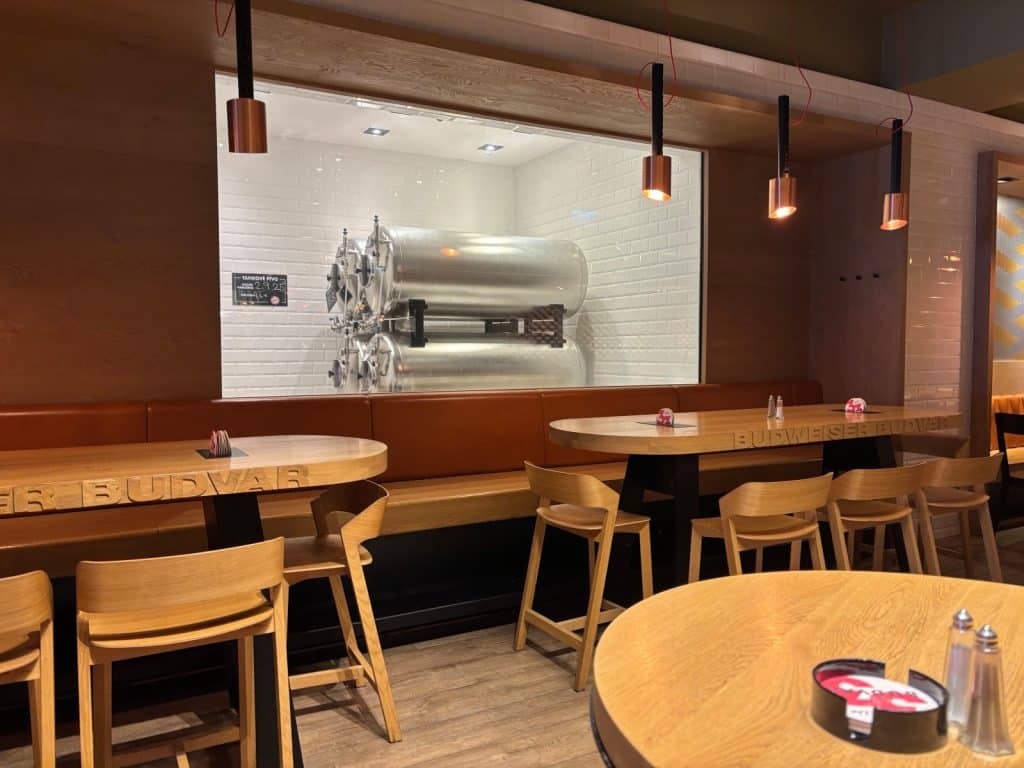 Ein modern gestalteter Gastraum im Budweiser Budvar Restaurant mit Holztischen, Stühlen und einer Glasfront, die den Blick auf große, silberne Brauereitanks freigibt.