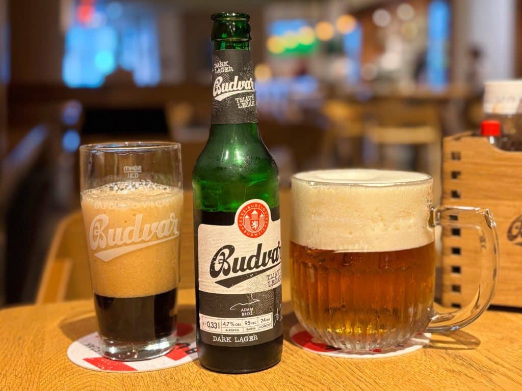 Eine Flasche Budvar Dark Lager steht in einem Restaurant zwischen einem Glas dunklem Bier und einem großen Krug hellem Bier auf einem Holztisch.