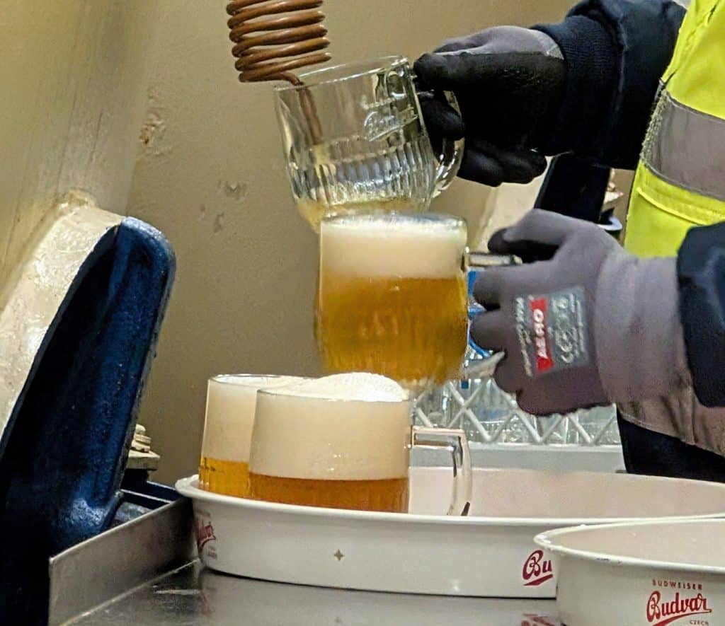 Ein Mitarbeiter in Arbeitskleidung zapft bei einer Brauereiführung in der Budweiser Budvar Brauerei frisches Bier aus einer kupfernen Kühlschlange in gläserne Krüge.