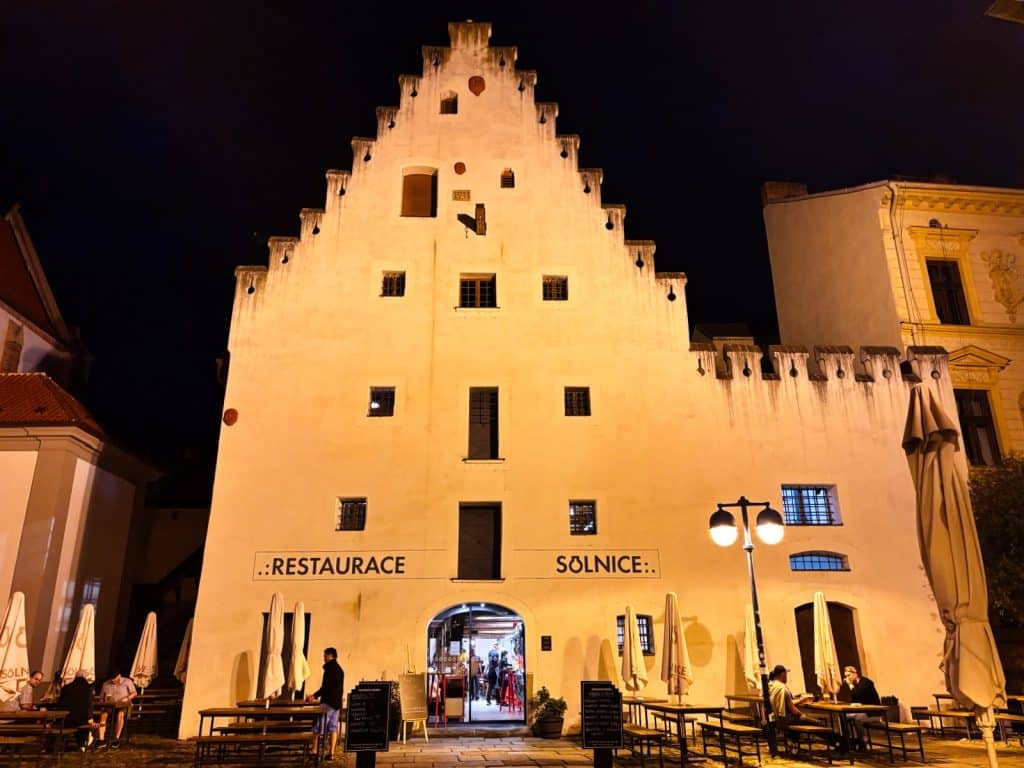 Das beleuchtete historische Gebäude des Restaurants Solnice in Budweis bei Nacht mit seiner markanten Treppengiebelfassade und einem Außenbereich mit Gästen an Holztischen.