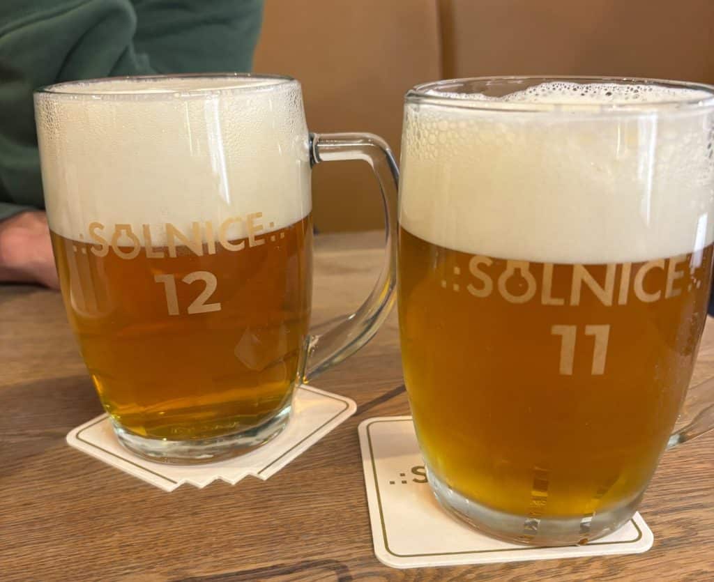 Zwei Gläser frisch gezapftes Bier der Brauerei Solnice in Budweis stehen mit einer ausgeprägten Schaumkrone auf Untersetzern auf einem Holztisch. Die Gläser tragen die Aufschriften „Solnice 12“ und „Solnice 11“, was auf die unterschiedlichen Stammwürzegehalte hinweist.