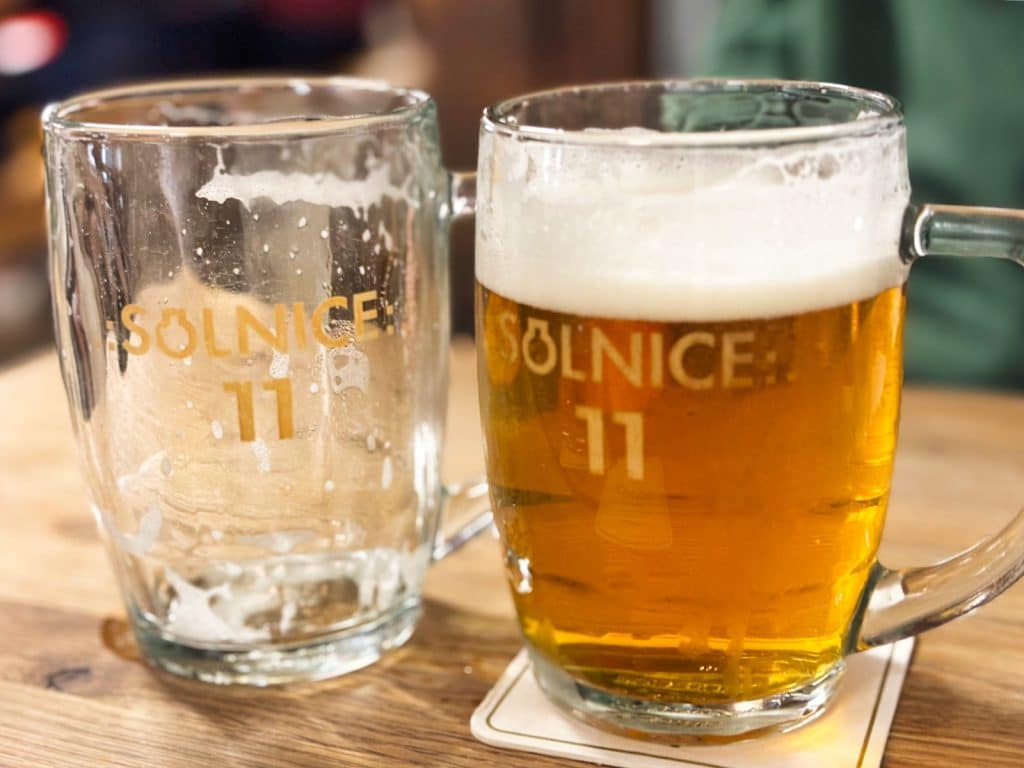 Zwei Bierkrüge der Brauerei Solnice in Budweis stehen auf einem Holztisch, wobei der linke Krug leer ist und der rechte mit hellem Bier und einer Schaumkrone gefüllt ist.