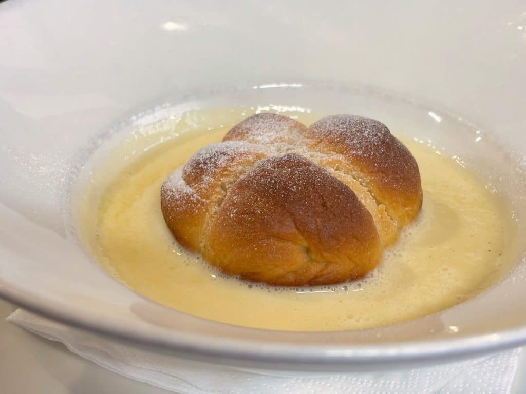 Ein goldbraun gebackener Hefekloß, bestäubt mit Puderzucker, ruht in einer hellen Vanillesauce in einem weißen, tiefen Teller im Restaurant Solnice in Budweis.