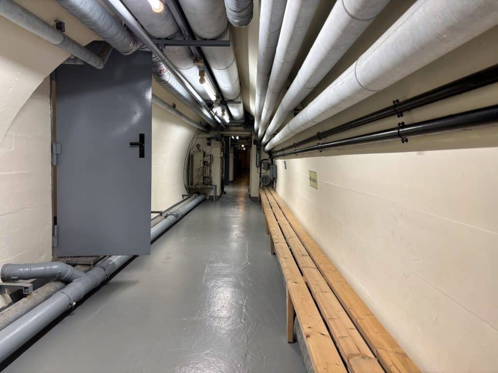 Ein langer, schmaler Gang im öffentlichen Schutzraum des Bundesbank-Bunkers in Cochem mit einer massiven grauen Stahltür, hölzernen Sitzbänken an der Wand und zahlreichen Versorgungsrohren an der Decke.