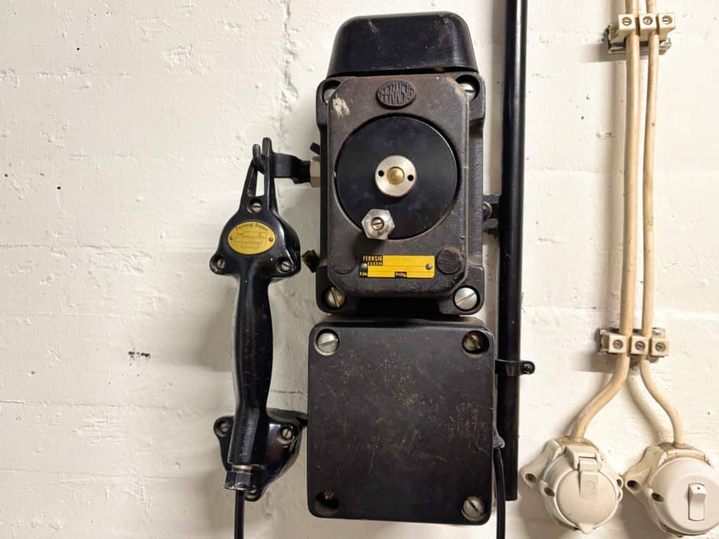 Ein robustes, schwarzes Wandtelefon aus Metall mit einem separaten Hörer an einer Halterung, installiert an einer weißen Wand im ehemaligen Bundesbank-Bunker in Cochem.