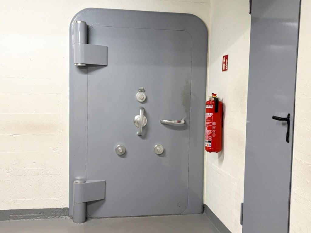 Eine massive, graue Stahltür mit großen Scharnieren und mehreren Verriegelungsmechanismen bildet den Tresoreingang im Bundesbank-Bunker in Cochem, neben dem ein roter Feuerlöscher an der weißen Wand hängt.
