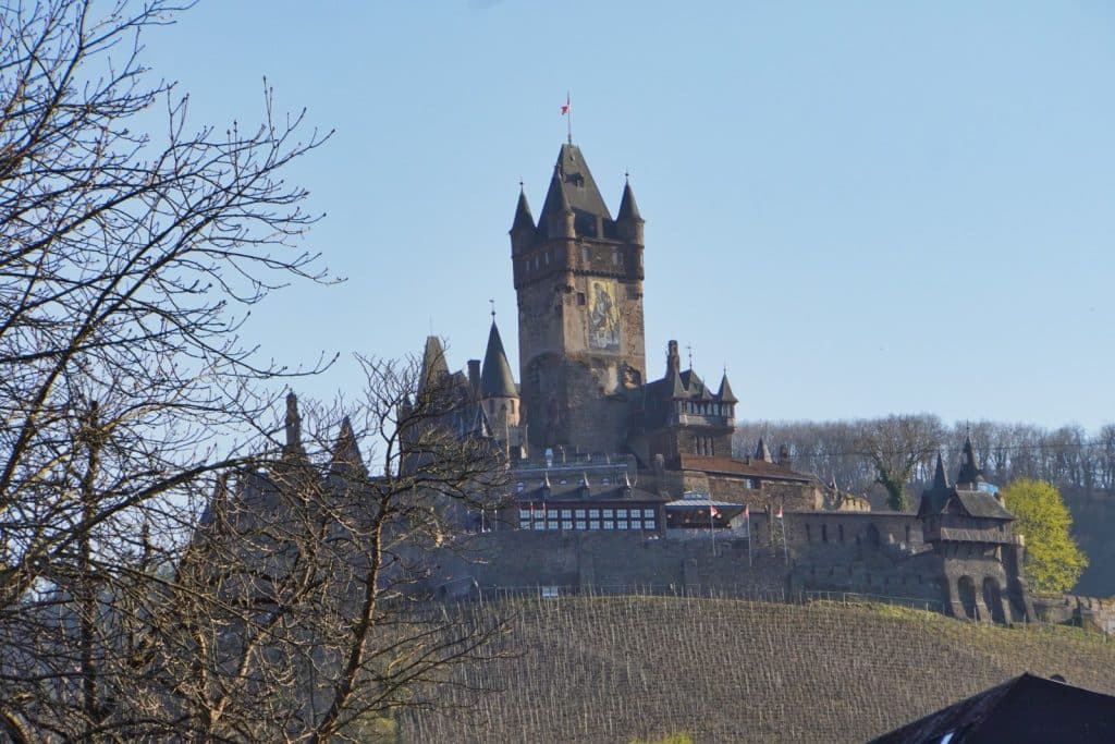 Eine Nahaufnahme der Reichsburg Cochem, die auf einem von Weinreben bewachsenen Hügel thront und von kahlen Bäumen im Vordergrund eingerahmt wird.