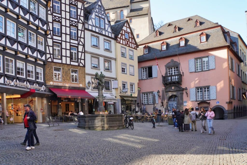 Der historische Marktplatz in Cochem mit seinen charakteristischen Fachwerkhäusern, einem zentralen Brunnen und Passanten auf dem Kopfsteinpflaster unter blauem Himmel.