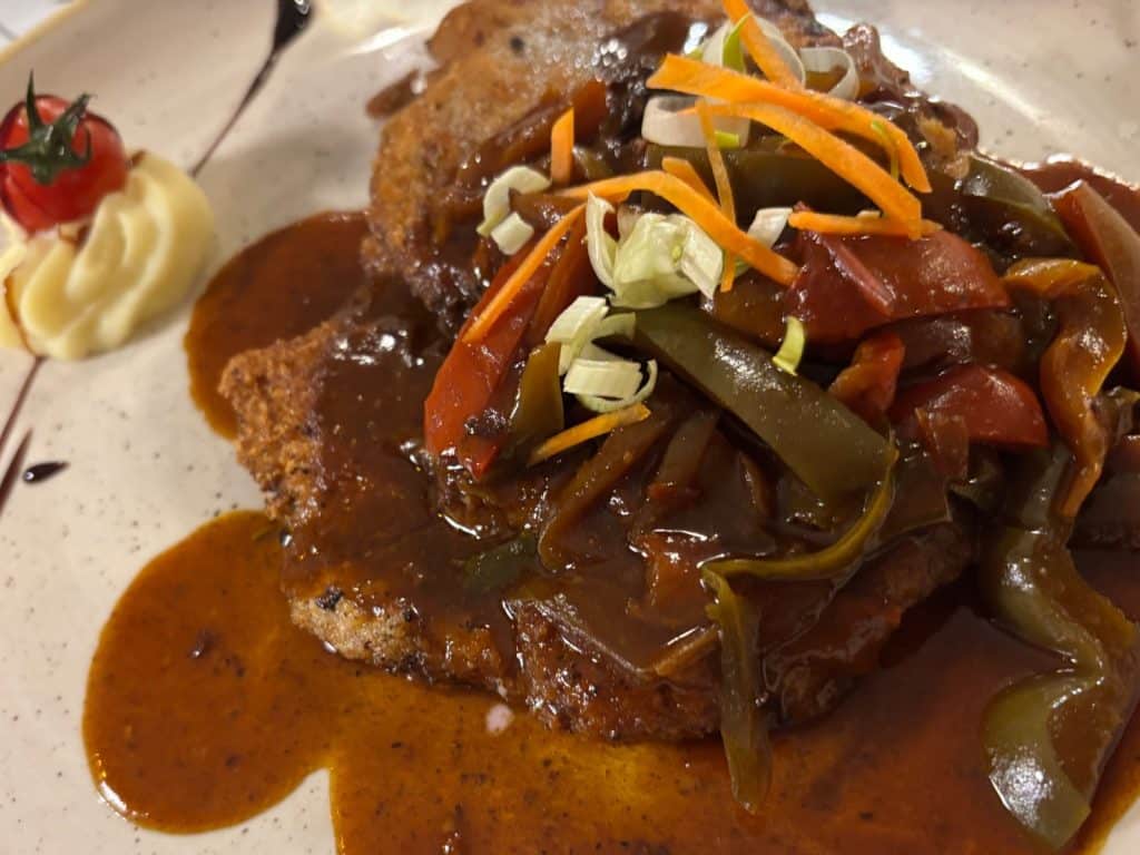 Ein im Restaurant Onkel Willi in Cochem serviertes Fleischgericht, garniert mit einer dunklen Sauce, geschmorten Paprikastreifen, Karottenstiften und Frühlingszwiebeln, serviert neben einem Klecks Kartoffelpüree mit einer Kirschtomate.