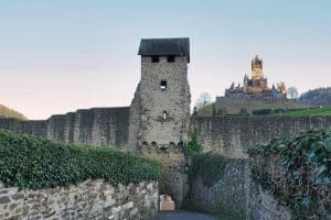 Ein steinerner Wachturm der historischen Stadtmauer von Cochem ragt über eine schmale Gasse empor, während im Hintergrund die Reichsburg Cochem auf einem Hügel thront.