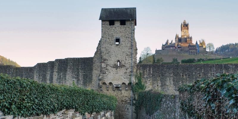 Ein steinerner Wachturm der historischen Stadtmauer von Cochem ragt über eine schmale Gasse empor, während im Hintergrund die Reichsburg Cochem auf einem Hügel thront.