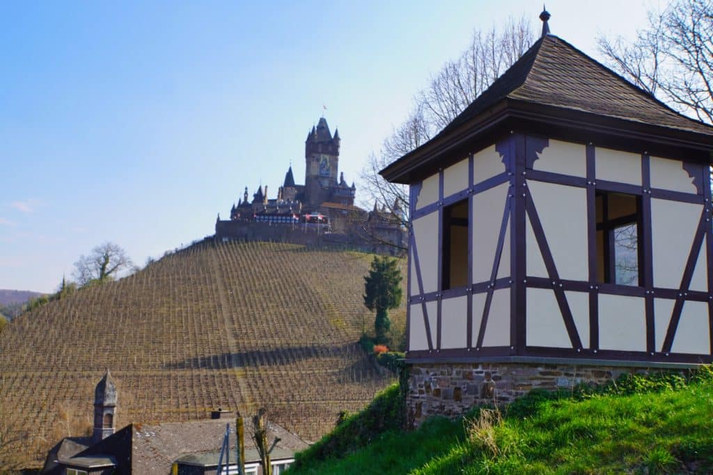 Blick von einem Aussichtspunkt in Cochem auf die Reichsburg, die über einem steilen Weinberg thront, mit einem Fachwerk-Pavillon im Vordergrund.