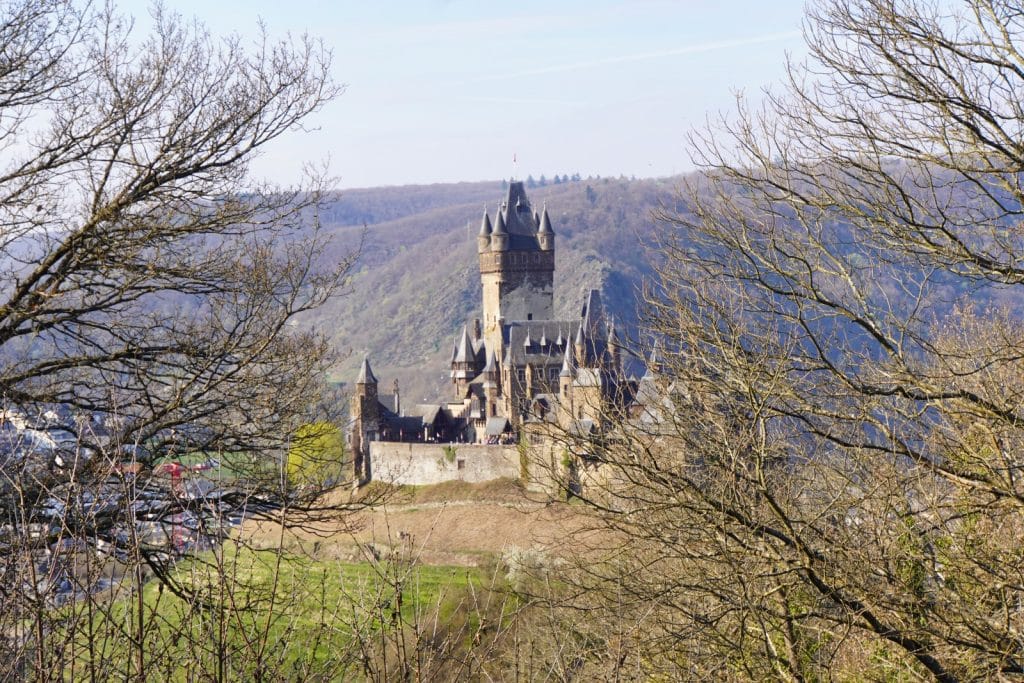 Blick durch kahle Baumzweige auf die thronende Reichsburg Cochem, die auf einem Hügel über dem Moseltal vor einer bewaldeten Bergkulisse liegt.