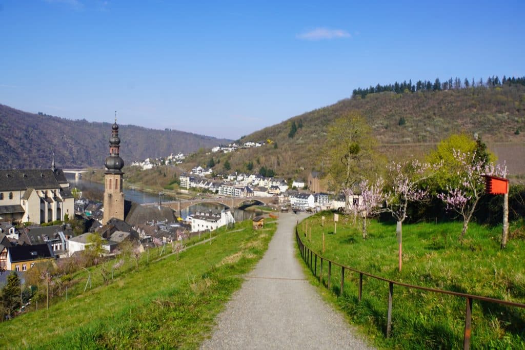 Ein geschotterter Wanderweg führt einen grünen Hang hinunter mit Blick auf die Stadt Cochem, die Moselbrücke und die markante St. Martin Kirche vor bewaldeten Hügeln.