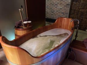 Eine leere, hölzerne Badewanne im Pilsen Bier Spa ist mit schäumendem Badewasser gefüllt, während daneben ein Holzfass mit Zapfhahn und zwei frisch gezapften Gläsern Bier steht.