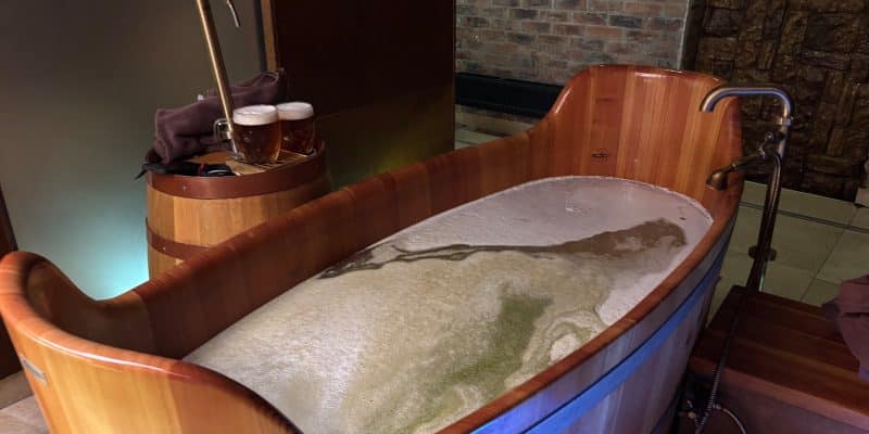 Eine leere, hölzerne Badewanne im Pilsen Bier Spa ist mit schäumendem Badewasser gefüllt, während daneben ein Holzfass mit Zapfhahn und zwei frisch gezapften Gläsern Bier steht.