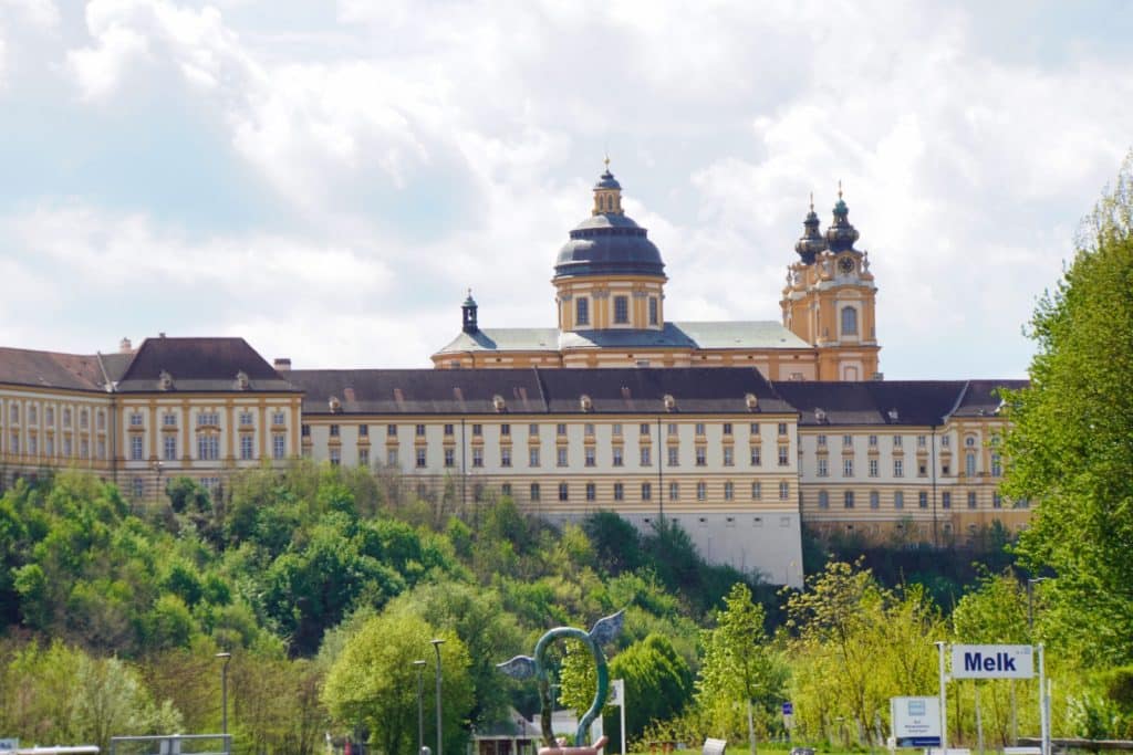 Das imposante, gelb-weiße Barockgebäude des Stift Melk thront auf einem bewaldeten Hügel über der Schiffsanlegestelle, im Vordergrund ist ein Schild mit der Aufschrift „Melk“ zu sehen.