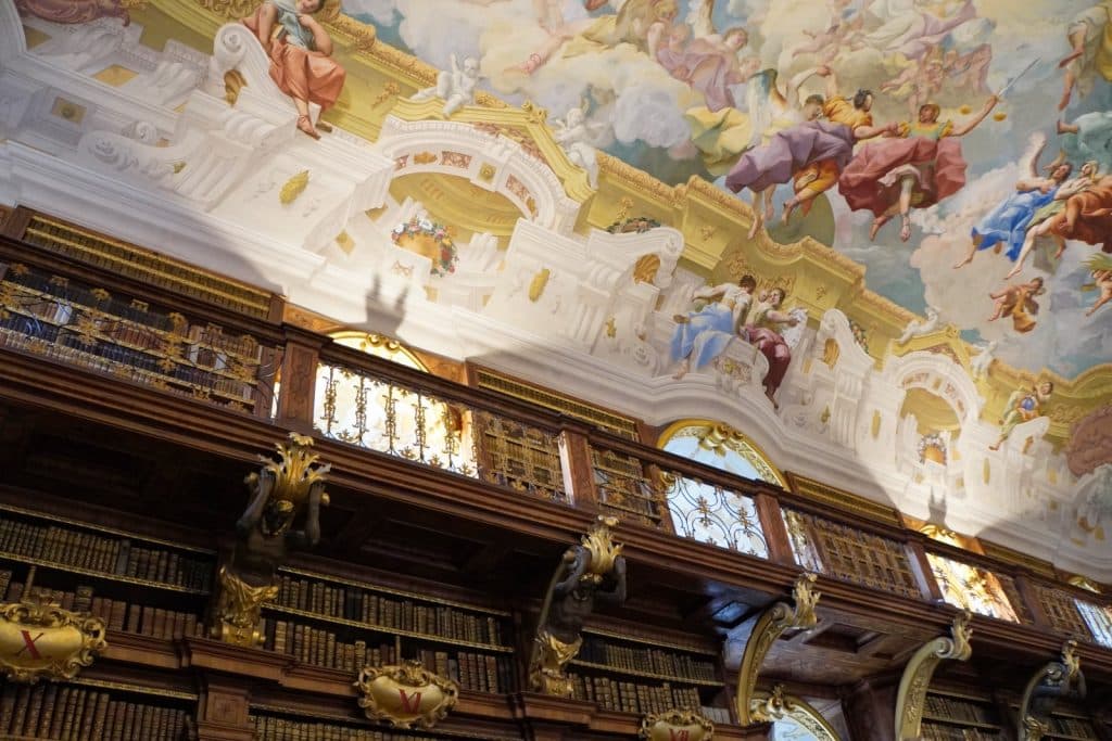 Blick von unten auf die prunkvolle Decke der Bibliothek im Stift Melk, die ein farbenfrohes barockes Deckenfresko mit zahlreichen Figuren in einer Himmelsdarstellung über reich verzierten, dunklen Holzregalen voller alter Bücher zeigt.