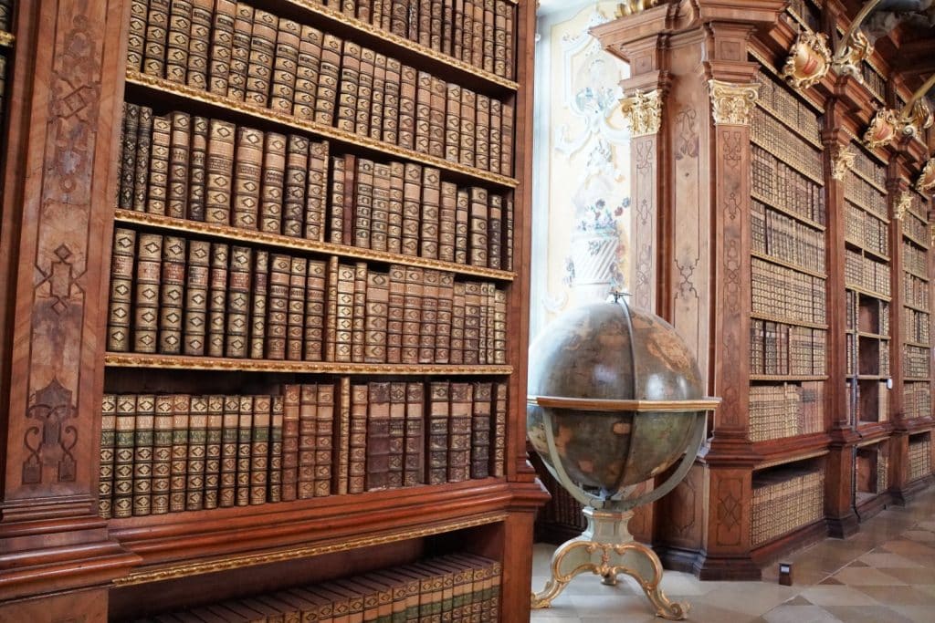 Prunkvolle Holzregale voller antiker, ledergebundener Bücher in der Bibliothek von Stift Melk, neben denen ein großer historischer Globus auf einem verzierten Standfuß steht.