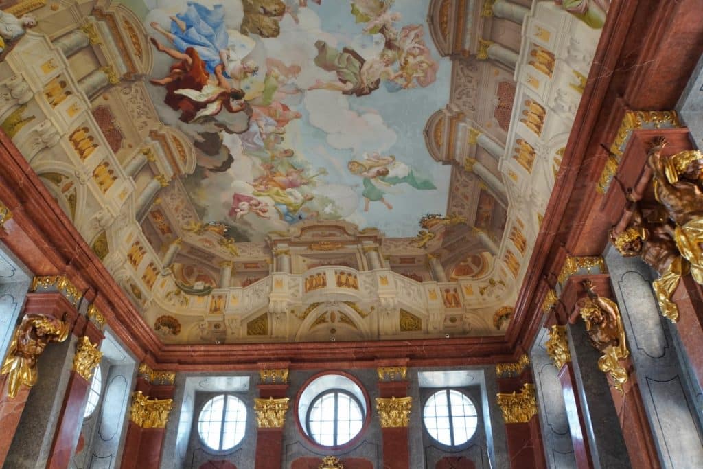 Blick nach oben auf das prunkvolle Deckenfresko im Marmorsaal des Stiftes Melk, das eine himmlische Szene mit mythologischen Figuren umrahmt von illusionistischer Architektur zeigt. Die prachtvolle Raumgestaltung wird durch rötliche Marmorsäulen, goldene Kapitelle und monumentale Skulpturen ergänzt.