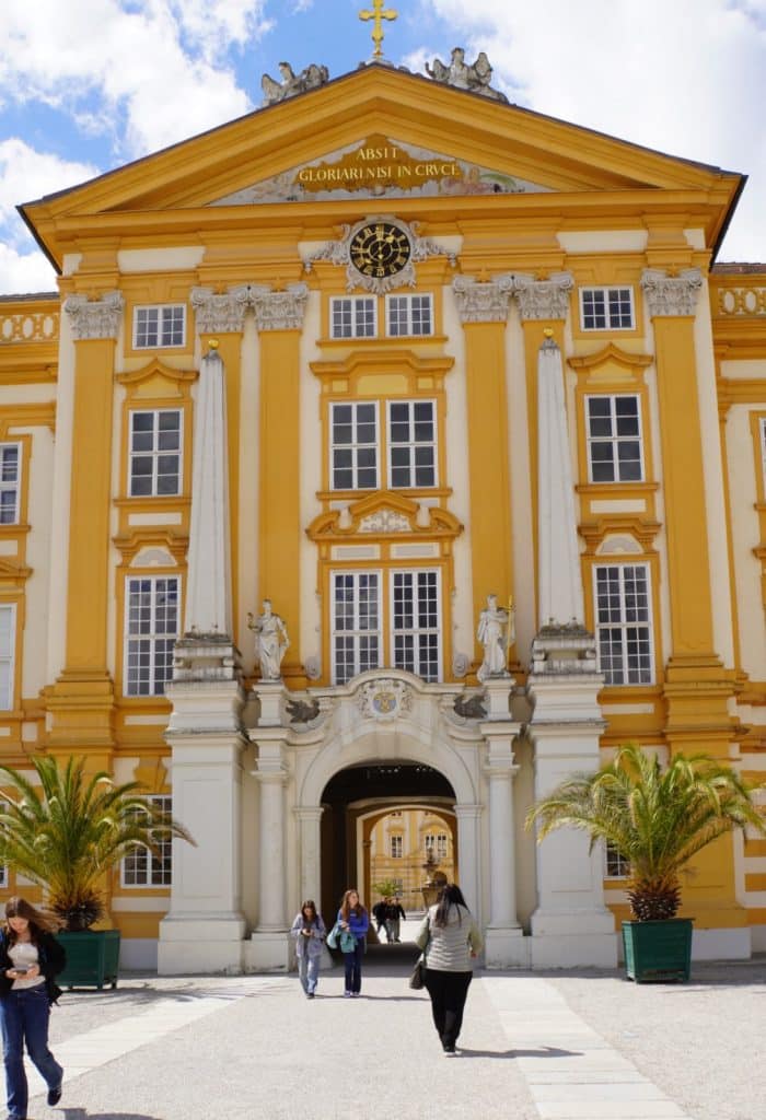 Die markante, gelb-weiße Barockfassade des Eingangsportals von Stift Melk mit einem zentralen Torbogen, Statuen und einer goldenen Inschrift unter dem Giebel. Im Vordergrund gehen Besucher über den gepflasterten Vorplatz, der von Palmen in Kübeln flankiert wird.